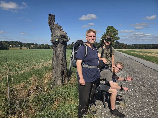Drei Wanderer mit Rucksäcken posieren lächelnd an einem Baumstamm am Straßenrand. Der Baumstamm trägt ein gelbes Schild mit einer Aufschrift. Im Hintergrund erstreckt sich eine weite, grüne Landschaft unter einem blauen Himmel mit einigen Wolken.