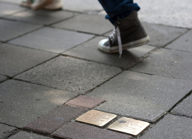 Neben zwei goldenen Stolpersteinen geht eine Person mit Turnschuhen vorbei, von der man nur die Füße sieht.