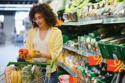 Eine Frau mit lockigen Haaren und Brille trägt ein gelbes Hemd über einem weißen Oberteil und steht an einem Supermarktregal. Sie hält eine rote und eine gelbe Paprika in den Händen und betrachtet sie aufmerksam. Im Einkaufswagen liegen verschiedene frische Lebensmittel, darunter Brot und Gemüse. Die Regale im Hintergrund sind mit verpacktem Obst und Gemüse gefüllt.