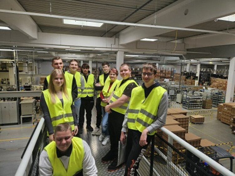 Eine Gruppe von neun jungen Erwachsenen trägt gelbe Warnwesten und steht auf einer Galerieebene in einer modernen Industriehalle. Sie posieren lächelnd für ein Gruppenfoto. Im Hintergrund sind Maschinen, Regale mit Kisten und Produktionsanlagen zu sehen, was auf eine Werksbesichtigung oder einen betrieblichen Rundgang hinweist.