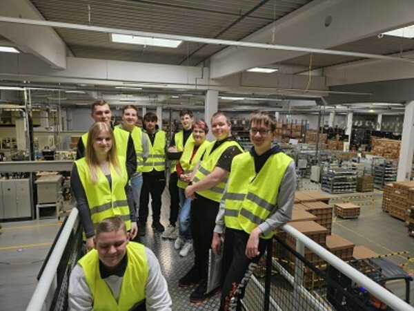 Eine Gruppe von neun jungen Erwachsenen trägt gelbe Warnwesten und steht auf einer Galerieebene in einer modernen Industriehalle. Sie posieren lächelnd für ein Gruppenfoto. Im Hintergrund sind Maschinen, Regale mit Kisten und Produktionsanlagen zu sehen, was auf eine Werksbesichtigung oder einen betrieblichen Rundgang hinweist.