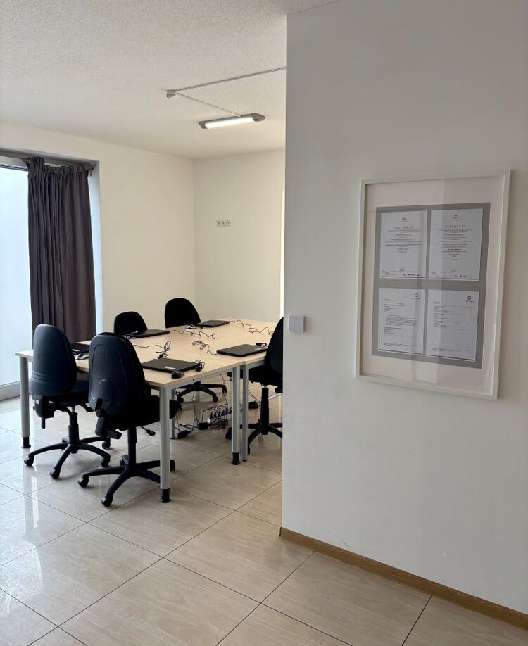 Ein kleinerer Besprechungs- oder Schulungsraum mit einem Tisch in U-Form, an dem mehrere Bürostühle mit Laptops und Kabeln platziert sind. An der Wand hängt ein Rahmen mit mehreren Dokumenten oder Zertifikaten. Der Raum ist hell, mit großen Fenstern und einem glatten Bodenbelag, und wirkt einladend und funktional.