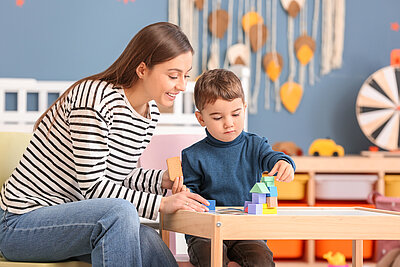 Eine junge Erzieherin betreut einen kleinen Jungen, der mit Bauklötzen spielt. Sie trägt ein geringeltes Oberteil und hält einen Keks in der Hand. Der Junge betrachtet konzentriert sein Bauwerk. Im Hintergrund befindet sich weiteres Spielzeug.