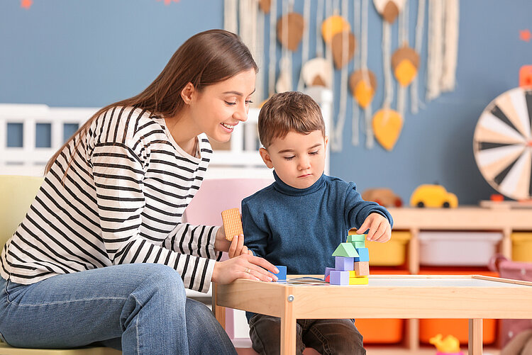 Eine junge Erzieherin betreut einen kleinen Jungen, der mit Bauklötzen spielt. Sie trägt ein geringeltes Oberteil und hält einen Keks in der Hand. Der Junge betrachtet konzentriert sein Bauwerk. Im Hintergrund befindet sich weiteres Spielzeug.