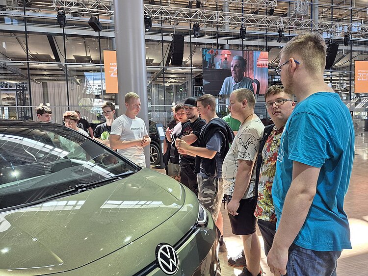 Zu sehen sind links mehrere junge Männer, die rechts ein dunkelgrün lackiertes Auto von VW anschauen.