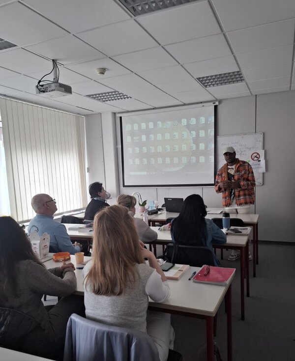 Ein männlicher Teilnehmender mit weißer Schirmmütze und orange-braun-karriertem Holzfällerhemd steht vorn am Whiteboard und trägt seinen Mitteilnehmenden des B2-Sprachkurses seine Präsentation vor.