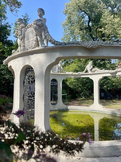 Runder Pavillon aus hellem Stein mit offenen Bögen und verzierten Metallgittern. Auf dem Dach stehen mehrere Skulpturen von Kindern, die Girlanden halten. Im Inneren befindet sich ein flacher, runder Teich mit grünem Wasser. Im Vordergrund sind unscharfe Blüten zu sehen, im Hintergrund hohe Bäume und blauer Himmel.