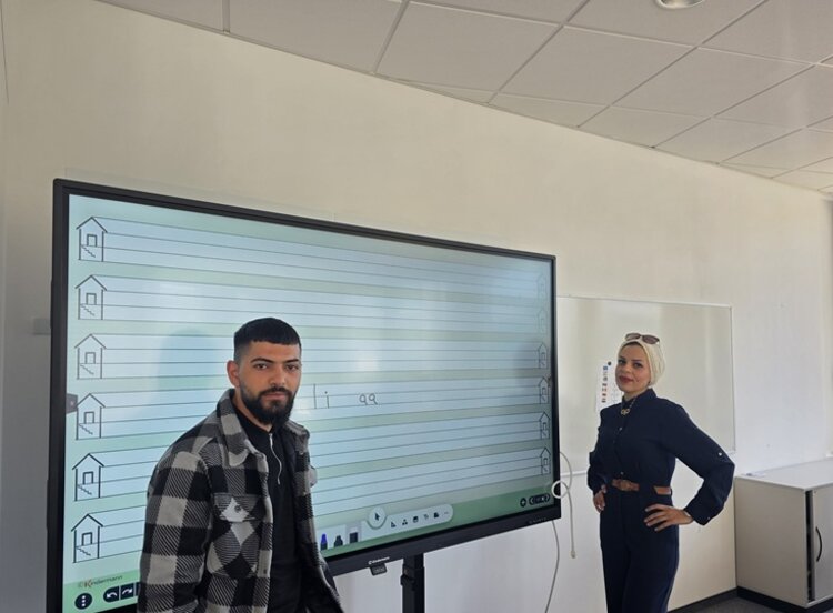 Ein männlicher Teilnehmer des Integrationskurses an den Euro-Schulen Dresden steht, gemeinsam mit der ehemaligen Teilnehmerin, die als Gast den Kurs besuchte, im Unterrichtsraum vor dem großen interaktiven Whiteboard.