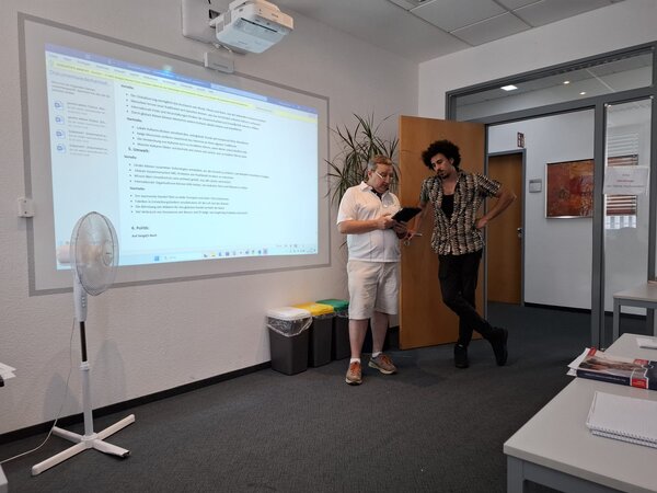 Ein Dozent steht gemeinsam mit einem Kursteilnehmer vor einer Präsentation, die auf eine Leinwand projiziert wird. Die beiden unterhalten sich, während auf der Leinwand Informationen zum Thema Globalisierung angezeigt werden.