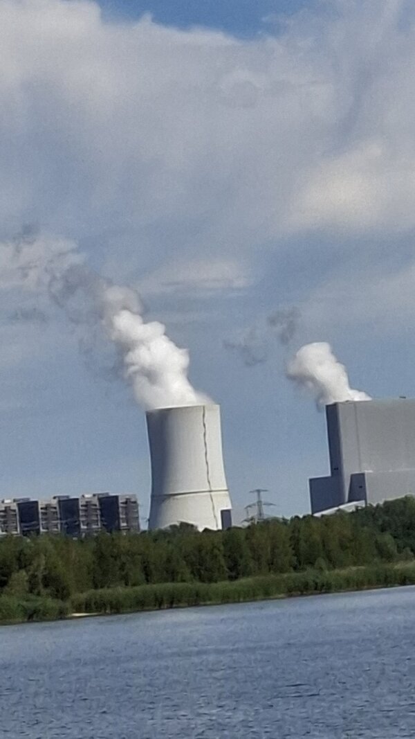 Blick über ein Gewässer auf ein Kraftwerk, aus dessen Kühlturm und Industriegebäude weißer Wasserdampf aufsteigt.