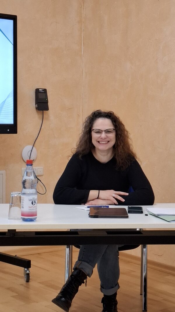 Die Referentin sitzt lächelnd an einem weißen Tisch in einem Seminarraum der Euro-Schulen Hoyerswerda. Vor ihr liegen ein Tablet, ein Smartphone, Unterlagen und ein Stift. Auf dem Tisch steht zudem eine Wasserflasche und ein Glas. Die Frau trägt eine schwarze Brille, ein schwarzes Oberteil, graue Jeans und schwarze Stiefel. Im Hintergrund ist eine beige strukturiert gestaltete Wand sowie ein Bildschirmrand sichtbar. Das Foto entstand im Rahmen einer Inhouse-Weiterbildung für Mitarbeitende zum Thema Kommunikation mit der Generation Z und zeigt eine freundliche, konzentrierte Seminaratmosphäre.