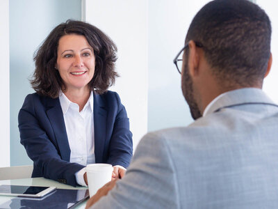 Eine Frau und ein Mann sitzen sich an einem Tisch gegenüber und unterhalten sich. Die Frau trägt einen dunkelblauen Blazer und ein weißes Hemd, während der Mann ein hellgraues Hemd trägt. Sie scheinen in ein Gespräch vertieft zu sein, möglicherweise in einem beruflichen oder formellen Kontext.