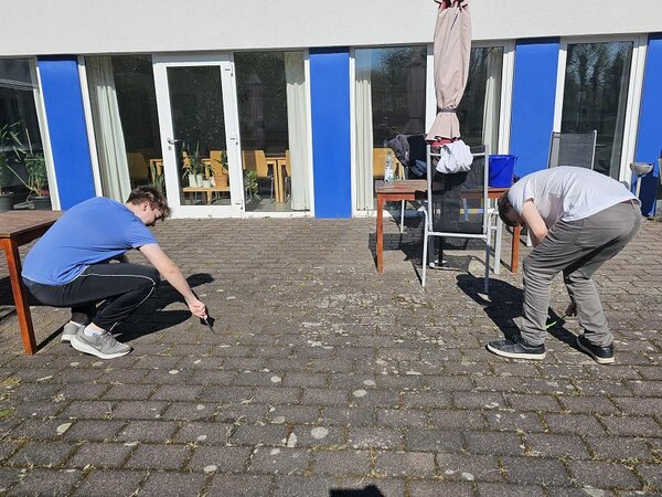 Zwei Personen reinigen gemeinsam den Boden einer Terrasse mit Unkrautkratzern. Im Hintergrund sind Tische, Stühle. ein Sonnenschirm und ein Gebäude mit großen Fenstern und blauen Wandteilen zu sehen.