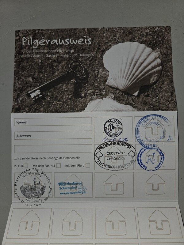 Ein Pilgerausweis für den Ökumenischen Pilgerweg durch Sachsen, Sachsen-Anhalt und Thüringen. Der Ausweis zeigt ein Bild mit einem Schlüssel und einer Muschel auf sandigem Untergrund. Es gibt Felder für Stempel und Unterschriften sowie Informationen auf Deutsch.