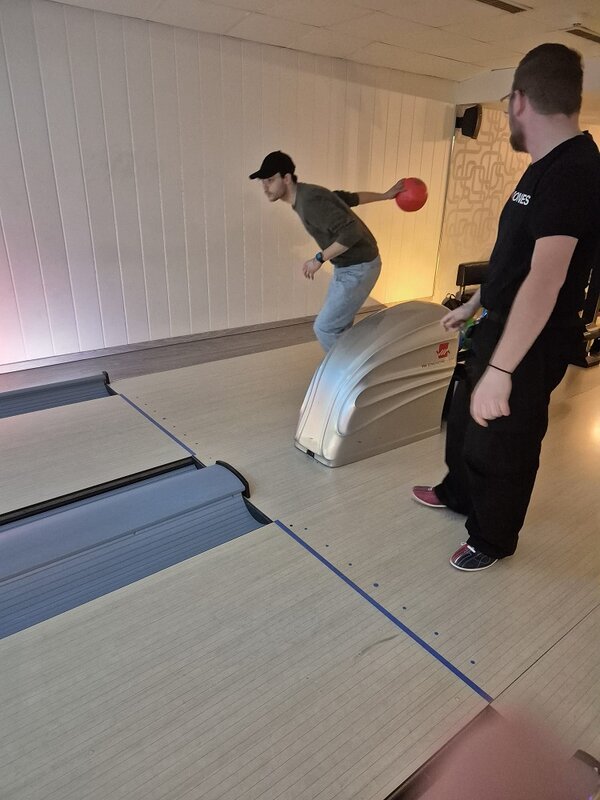 Ein Teilnehmer mit Kappe holt zum Wurf mit einer roten Bowlingkugel aus, während ein weiterer Teilnehmer am Bahnanfang danebensteht und zuschaut.