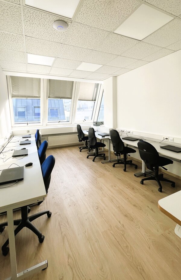 Schmaler Computerraum mit zwei Reihen Arbeitsplätzen: weiße Tische mit Laptops und Bürostühlen entlang der beiden Wände, vorne große schräge Fenster.