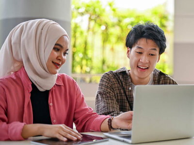 Zwei Personen sitzen an einem Tisch und arbeiten gemeinsam an einem Laptop. Eine Person in einem rosa Hemd und heller Kopfbedeckung zeigt auf den Bildschirm, während die andere Person den Laptop betrachtet. Auf dem Tisch liegen zusätzlich ein Tablet und Arbeitsmaterialien. Im Hintergrund sind helle Fenster und grüne Pflanzen zu sehen.