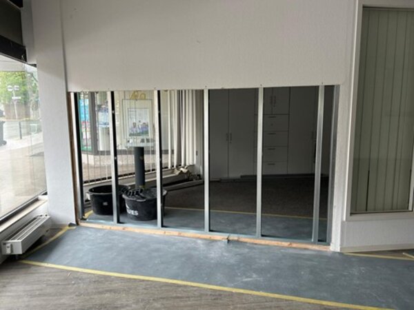 Blick auf eine Baustelle in einem Raum. Scheinbar wird eine zusätzliche Wand gebaut. Man sieht die Metallstreben und man kann noch in den anderen Raum blicken. Links sind große, bodentiefe Fenster und zwei große Eimer mit Bauschutt zu sehen.