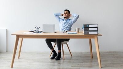 Ein lächelnder Mann sitzt entspannt auf einem Bürostuhl am Schreibtisch, vor einem Laptop, lehnt sich zurück und ist zufrieden mit seinem Job.