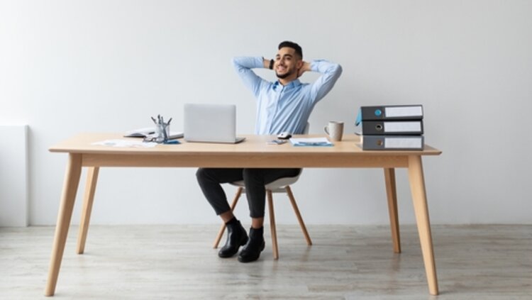 Ein lächelnder Mann sitzt entspannt auf einem Bürostuhl am Schreibtisch, vor einem Laptop, lehnt sich zurück und ist zufrieden mit seinem Job.