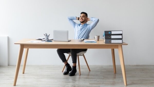 Ein lächelnder Mann sitzt entspannt auf einem Bürostuhl am Schreibtisch, vor einem Laptop, lehnt sich zurück und ist zufrieden mit seinem Job.