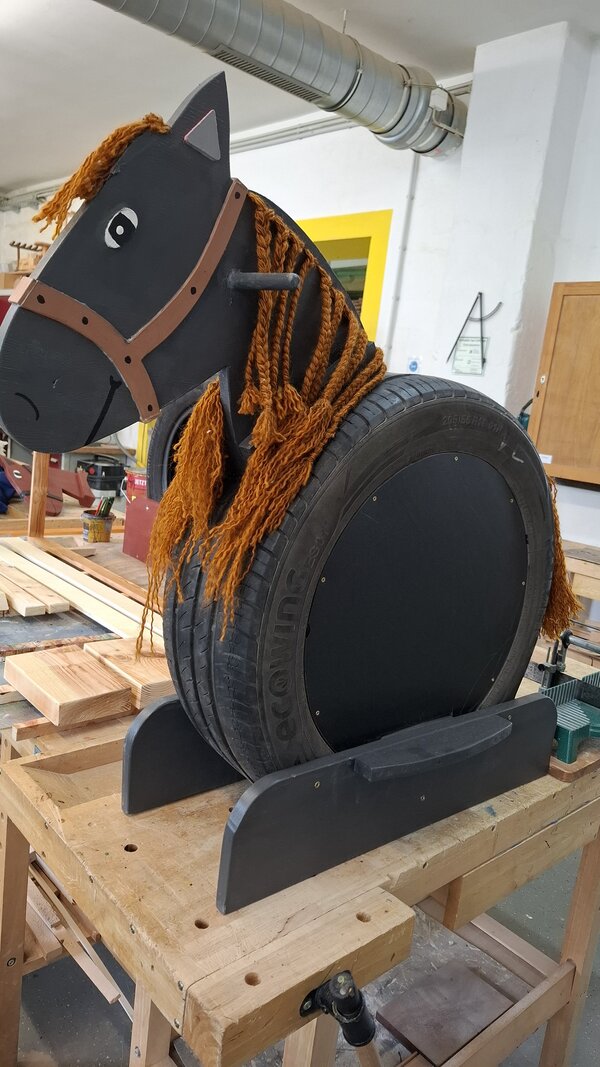 Auf dem Bild ist ein kleines aus Holz und mit schwarzer Farbe angemaltes Pferd mit goldener Kette zu sehen.