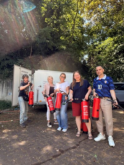 Fünf lachende Personen stehen im Freien vor einem weißen Anhänger und halten rote Feuerlöscher in den Händen. Über ihnen scheint Sonnenlicht durch dichte Bäume.
