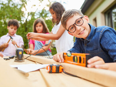 Ein Junge mit Brille und einem blauen Hemd sitzt an einem Tisch und hält eine orangefarbene Wasserwaage. Er lächelt in die Kamera. Im Hintergrund sind drei weitere Kinder, die an einem Bastelprojekt arbeiten. Ein Kind hält einen Hammer, während die anderen mit verschiedenen Werkzeugen und Materialien beschäftigt sind. Der Tisch ist mit Werkzeugen und Papier bedeckt, was auf eine kreative oder handwerkliche Aktivität hinweist.