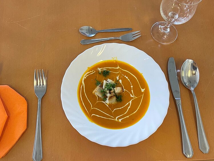 BRAFO-Projekt im Oktober – Herbst trifft Halloween! Ein weißer Teller mit Kürbissuppe steht auf dem Tisch, daneben Besteck, eine orangefarbene Serviette und ein Wasserglas. Die Suppe ist mit Croûtons, Kräutern und Sahne, die zu einem Spinnennetz auf ihr angerichtet ist.