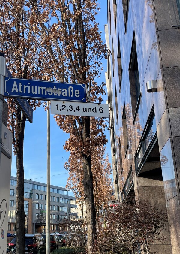 Straßenschild „Atriumstraße“ mit zusätzlichem weißen Hinweisschild „1,2,3,4 und 6“ vor Bäumen mit braunem Laub und umliegenden Bürogebäuden an einem sonnigen Tag.