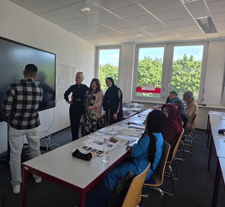 Die Teilnehmenden des Integrationskurses an den Euro-Schulen Dresden sitzen an Tischen, einige stehen vorn am Whiteboard mit der ehemaligen Teilnehmerin, die als Gast den Kurs besuchte, im Unterrichtsraum und posieren für ein Foto.