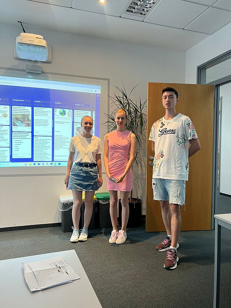 Heiße Köpfe, kühles Eis – Sprachkurse bei 35 Grad! Drei Kursteilnehmer stehen im Klassenraum vor einer digitalen Präsentation, die über einen Beamer an die Wand projiziert wird. Die Gruppe präsentiert ihr Ergebnis, im Hintergrund sind Pflanzen und die offene Tür zu sehen.