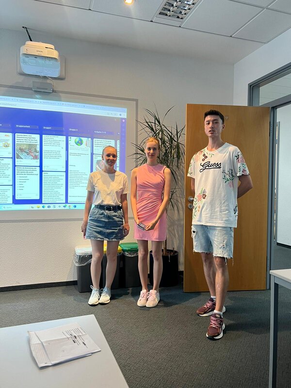 Drei Kursteilnehmer stehen im Klassenraum vor einer digitalen Präsentation, die über einen Beamer an die Wand projiziert wird. Die Gruppe präsentiert ihr Ergebnis, im Hintergrund sind Pflanzen und die offene Tür zu sehen.