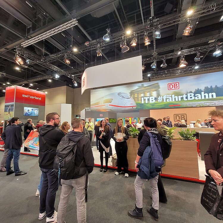 „BO auf Tour – Unternehmen laden ein“ auf der ITB 2025 Eine größere Gruppe von Menschen befindet sich an einem Messestand der Deutschen Bahn mit dem Slogan „ITB #fährtBahn“. Ein ICE-Zug ist auf einer großen digitalen Anzeige zu sehen, während Besucher in Gespräche vertieft sind. Im Hintergrund gibt es einen weiteren Stand für "Switzerland" mit Bildschirmen.