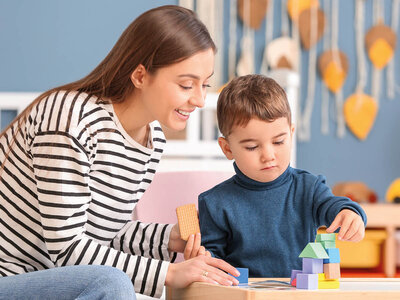 Eine angehende pädagogische Fachkraft unterstützt ein Kleinkind beim Bauen mit bunten Holzklötzen. Beide sitzen an einem kleinen Tisch in einem freundlich gestalteten Raum mit herbstlicher Dekoration.