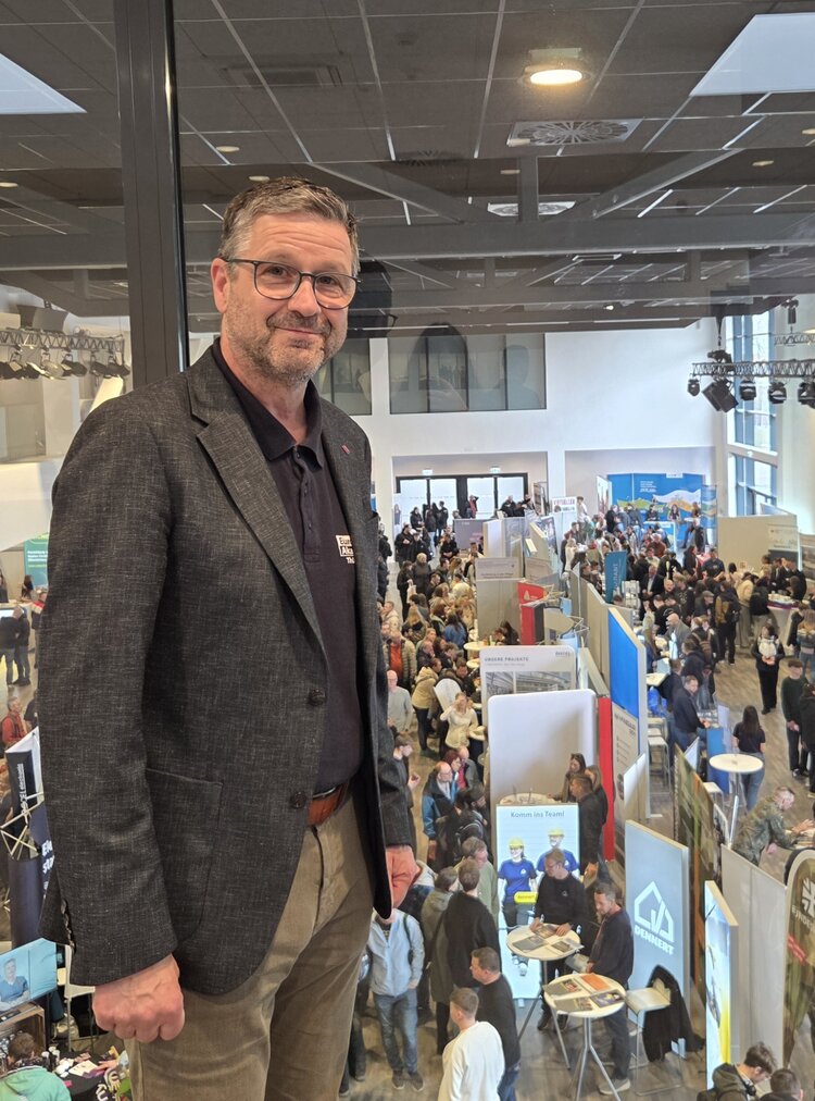 Im Vordergrund steht der Leiter Herr Schöneck in einem dunklen Sakko und einem dunkelblauen Poloshirt, er lächelt freundlich in Richtung Kamera und steht an einer Glasbrüstung. Im Hintergrund ist eine große Messehalle mit zahlreichen Messeständen und vielen Besuchern zu sehen. Die Stände sind mit verschiedenen Plakaten und Bannern ausgestattet, und die Halle ist hell beleuchtet. Die Decke zeigt ein Raster aus quadratischen Paneelen und mehrere Lichtspots.