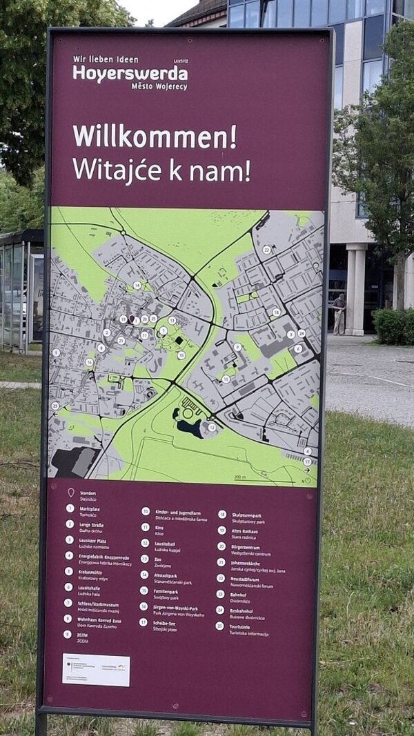 Eine dunkelrote Tafel mit einem Stadtplan von Hoyerswerda steht auf einer Wiese vor Gebäuden.