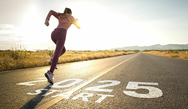 Eine Frau in Sportkleidung startet auf einer Landstraße ins Laufen. Auf dem Asphalt steht in großen weißen Buchstaben „2025 START“. Die Sonne geht am Horizont auf und beleuchtet die Szene warm, während sich die Straße in die Ferne erstreckt.