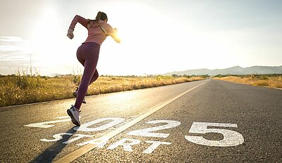 Eine Frau in Sportkleidung startet auf einer Landstraße ins Laufen. Auf dem Asphalt steht in großen weißen Buchstaben „2025 START“. Die Sonne geht am Horizont auf und beleuchtet die Szene warm, während sich die Straße in die Ferne erstreckt.