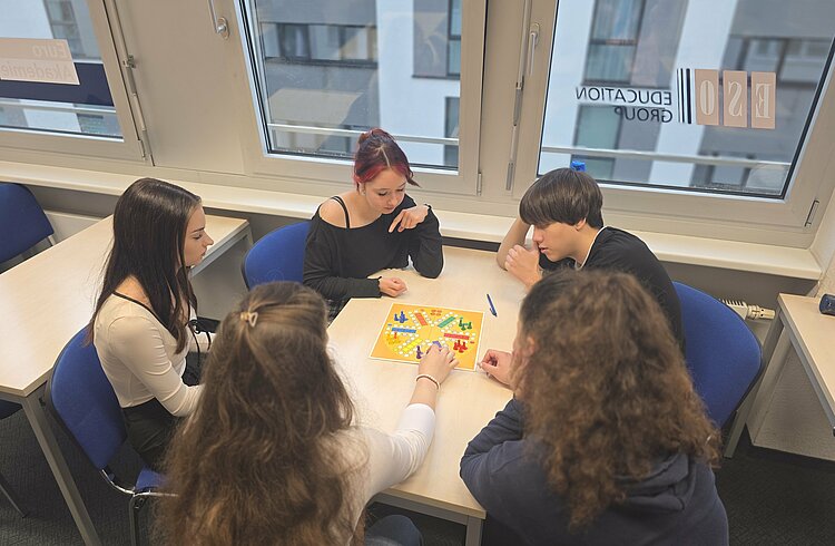 Fünf junge Erwachsene sitzen gemeinsam an einem Tisch und spielen „Mensch ärgere dich nicht“. Alle sind in das Gesellschaftsspiel vertieft.