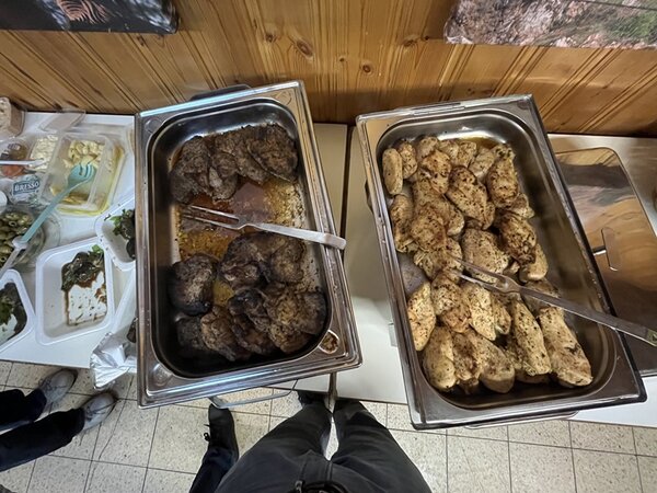 Zwei große Edelstahlbehälter stehen nebeneinander auf einem Buffet. Der linke Behälter enthält mehrere dunkle, gebratene Fleischstücke, der rechte Behälter ist mit helleren, ebenfalls gebratenen Fleischstücken gefüllt. Beide Behälter haben jeweils eine Metallzange zum Servieren. Im Hintergrund sind weitere Speisen in Kunststoffbehältern und ein hölzernes Wandpaneel sichtbar. Der Boden besteht aus hellen Fliesen.