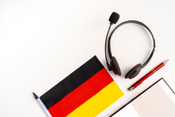 Auf einem weißen Hintergrund liegen eine Deutschlandflagge, ein Headset, ein Kugelschreiber und ein Schreibblock.
