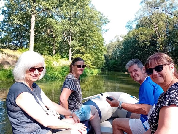 Drei Frauen mit Sonnenbrillen und ein Mann sitzen in einem Schlauchboot und fahren auf einem kleinen Fluss, an dessen Ufer verschiedene Bäume und Sträucher stehen.