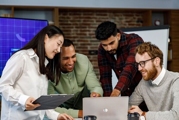 Ein Team junger multinationaler Geschäftsleute arbeitet gemeinsam an einem Laptop in einem modernen Büro mit Ziegelwänden und einem großen Bildschirm im Hintergrund.