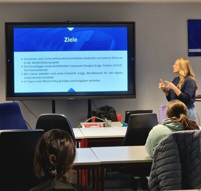 Eine Person steht vorne in einem Seminarraum und hält eine Präsentation. Auf dem großen Bildschirm hinter ihr steht das Wort "Ziele" und eine Aufzählung von Lernzielen auf Deutsch, darunter das Verständnis von Unternehmenskommunikation und Markenbildung, das Kennenlernen von Corporate Design-Grundlagen, das Erstellen von Entwürfen mit Canva, und das Gründen einer fiktiven Firma in Teams. Die Person präsentiert aktiv und deutet mit der Hand auf den Bildschirm. Im Vordergrund sitzen zwei Zuhörerinnen an Tischen und folgen der Präsentation.