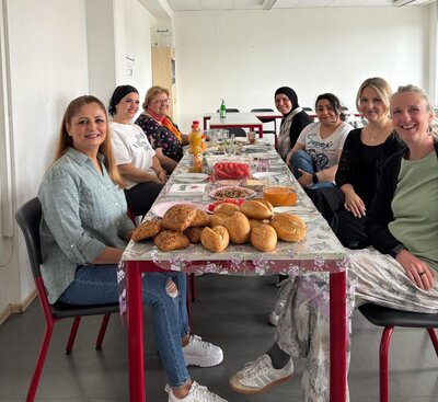 Eine Gruppe von sieben Frauen sitzt gemeinsam an einem langen Tisch in einem hellen Raum. Auf dem Tisch stehen verschiedene Speisen wie Brötchen, Obst, Snacks und Getränke. Die Frauen lächeln und wirken entspannt. Der Raum hat weiße Wände und rote Stühle.