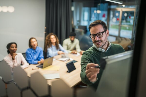 Ein Mann präsentiert in einem modernen Besprechungsraum Inhalte an einem Bildschirm, während vier Kolleg*innen an einem Tisch mit Laptops und Unterlagen zuhören.