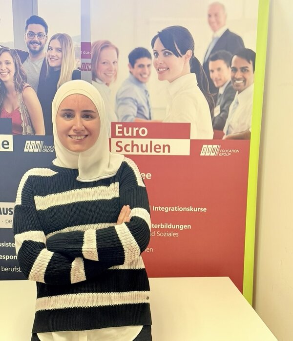 Eine junge Frau mit hellgrauem Kopftuch und gestreiftem Pullover steht mit verschränkten Armen vor einem Roll-up der Euro-Schulen und lächelt freundlich in die Kamera. Das Roll-up zeigt mehrere unscharf dargestellte Personen und trägt die Aufschrift 'Euro Schulen' sowie weitere Textzeilen. Die Farben des Plakats sind überwiegend rot, weiß und gelb. Im Hintergrund sind Logos und Textfelder zu sehen.