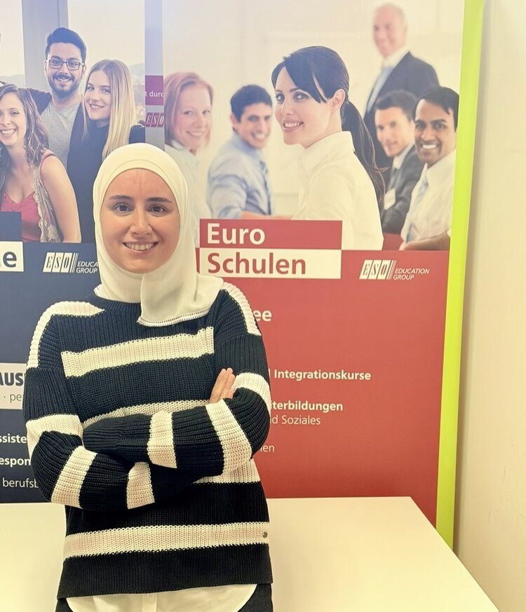 Eine junge Frau mit hellgrauem Kopftuch und gestreiftem Pullover steht mit verschränkten Armen vor einem Roll-up der Euro-Schulen und lächelt freundlich in die Kamera. Das Roll-up zeigt mehrere unscharf dargestellte Personen und trägt die Aufschrift 'Euro Schulen' sowie weitere Textzeilen. Die Farben des Plakats sind überwiegend rot, weiß und gelb. Im Hintergrund sind Logos und Textfelder zu sehen.