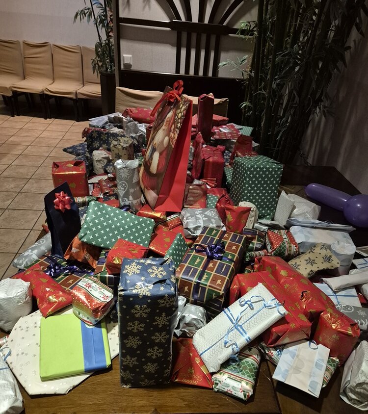 Ein großer Tisch, bedeckt mit einer Vielzahl unterschiedlich großer und farblich vielfältig verpackter Geschenke, die im Rahmen der Weihnachtsfeier der Euro-Schulen Rhein-Ruhr-Wupper GmbH arrangiert sind. Die Geschenkverpackungen zeigen verschiedene Muster, darunter Schneeflocken, Sterne, Karos und Schleifen in Farben wie Rot, Grün, Blau, Weiß und Gold. Einige Geschenke sind in Geschenkpapier eingewickelt, andere in Geschenktüten mit weihnachtlichen Motiven. Im Hintergrund sind Stühle an der Wand und Pflanzen zu sehen, die den Raum gemütlich wirken lassen. Die Szene vermittelt eine festliche Atmosphäre mit einem Fokus auf die zahlreichen Präsente, die auf dem Tisch liegen.